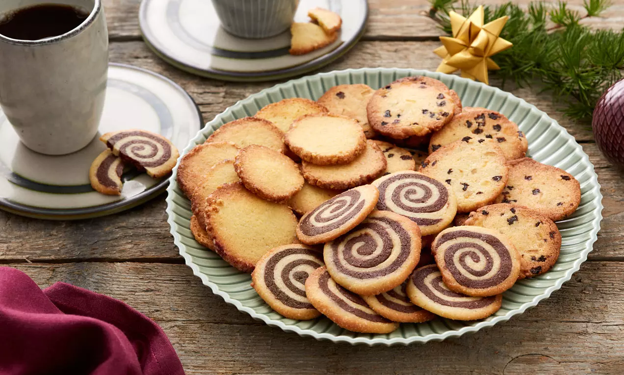 Småkager i gyldne nuancer med spiralform, chokoladeprikker og sprøde kanter anrettet på en lysegrøn tallerken ved siden af kaffe og julepynt