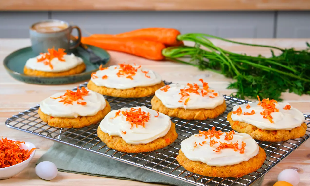 Lysebrune cookies med hvid frosting og revet orange gulerod på en metalrist, omgivet af friske gulerødder og et gråt krus