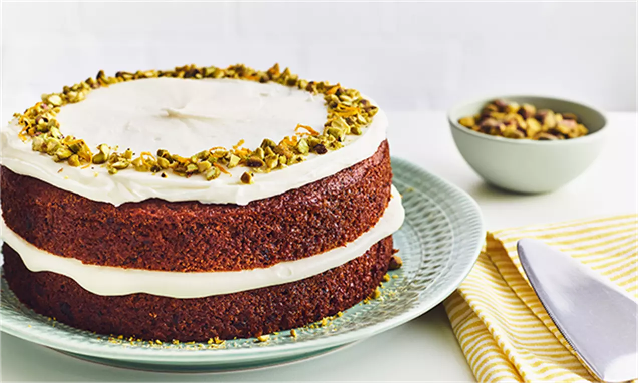 Vegansk gulerodskage med dyb orangebrun bund, hvid creme mellem lagene og på toppen, pyntet med grønne hakkede pistacienødder på en lysegrøn tallerken
