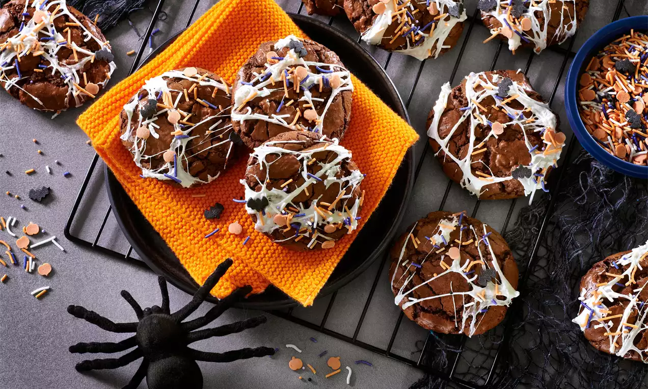 Mørkebrune cookies med ujævne marshmallow-striber og orange-blå pynt på en sort tallerken med orange serviet, omgivet af Halloween-dekoration