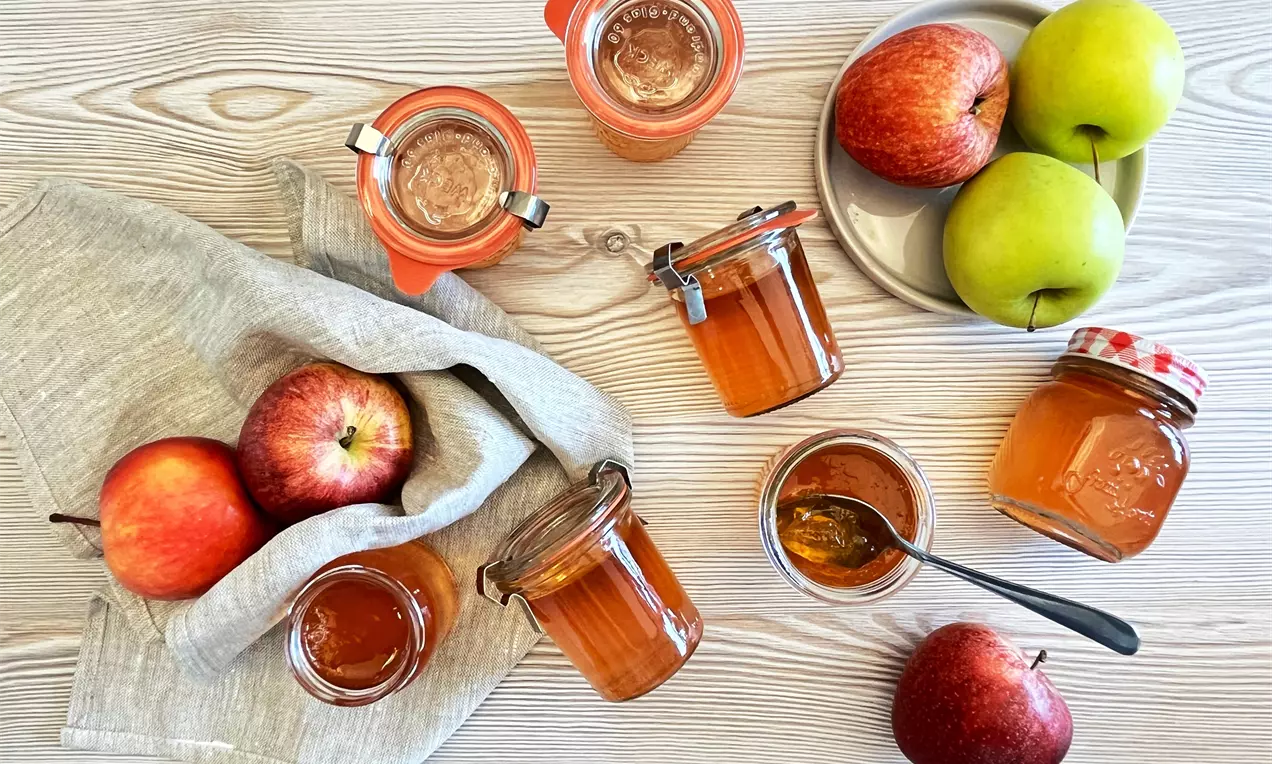 Klar orange æblegelé i gennemsigtige glas omgivet af røde og grønne æbler på lyst træbord med lysegråt klæde og en ske