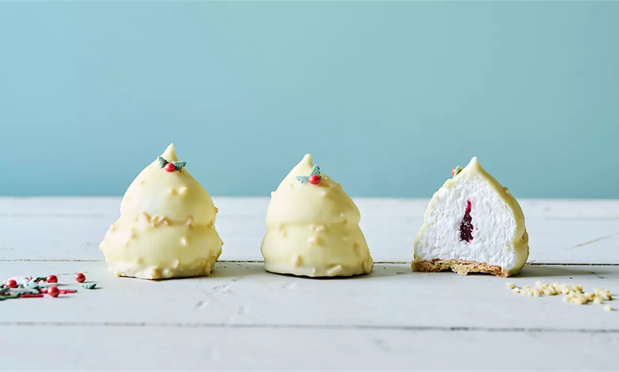Flødeboller med lys gul chokoladeoverflade pyntet med rødt og grønt krymmel, på en hvid, trætallerken mod lyseblå baggrund