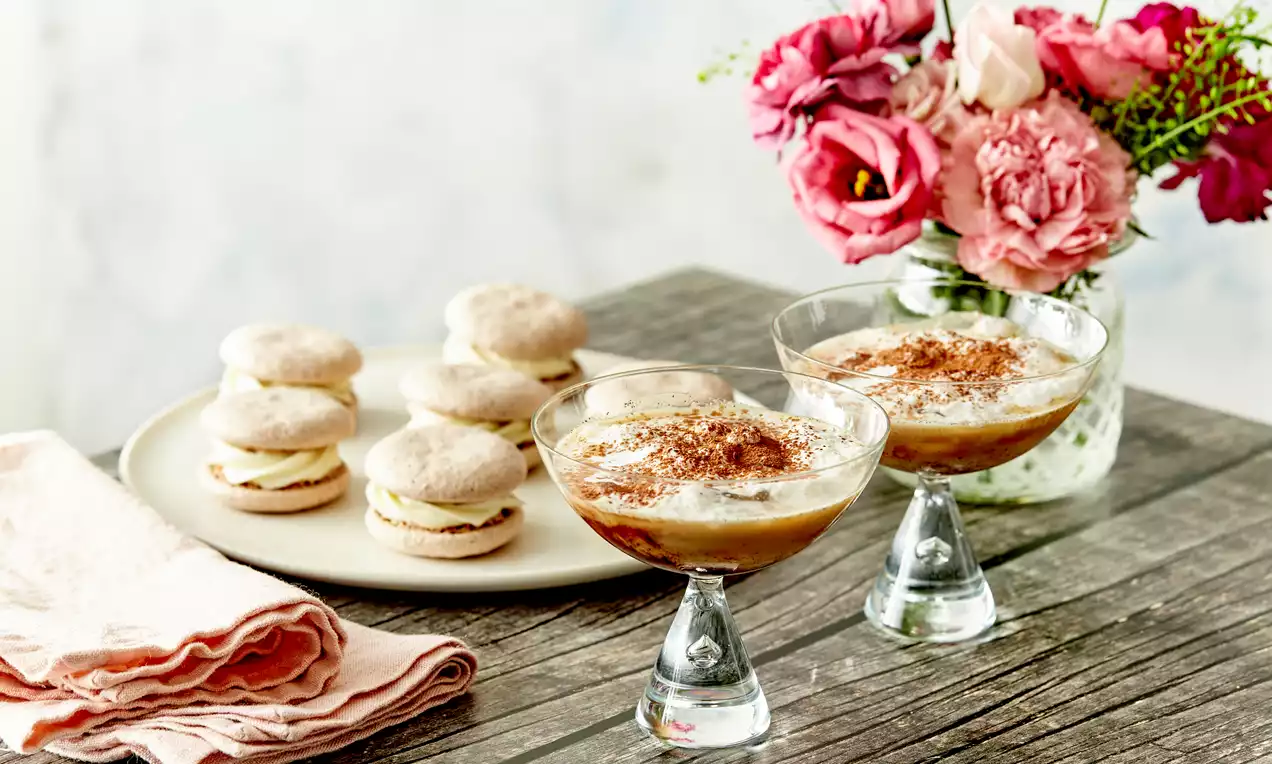 Tiramisu mocktails med cremet hvid skum og brun kakao på toppen serveret i klare glas ved siden af lyse macarons på fad og rosa blomster