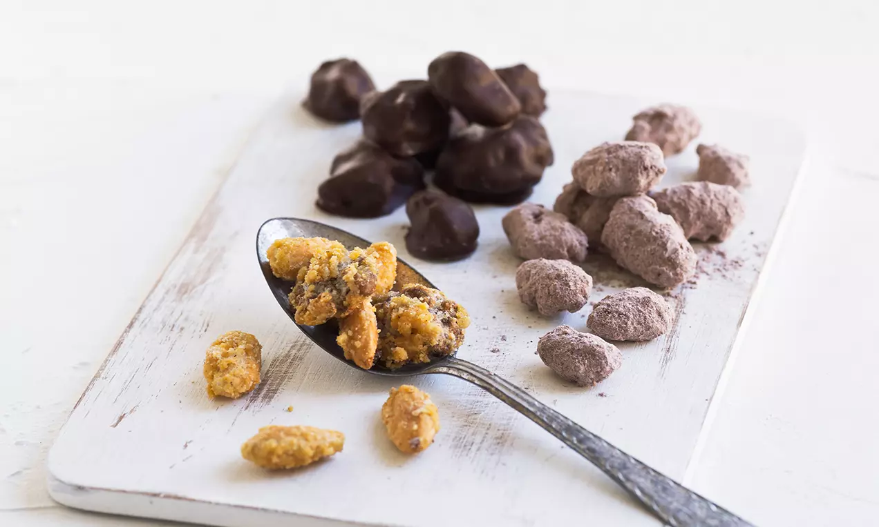 Ristede kakaonødder med mørk chokolade, lyse kryddernødder og karamelknas arrangeret på en hvid træplade med en metalske