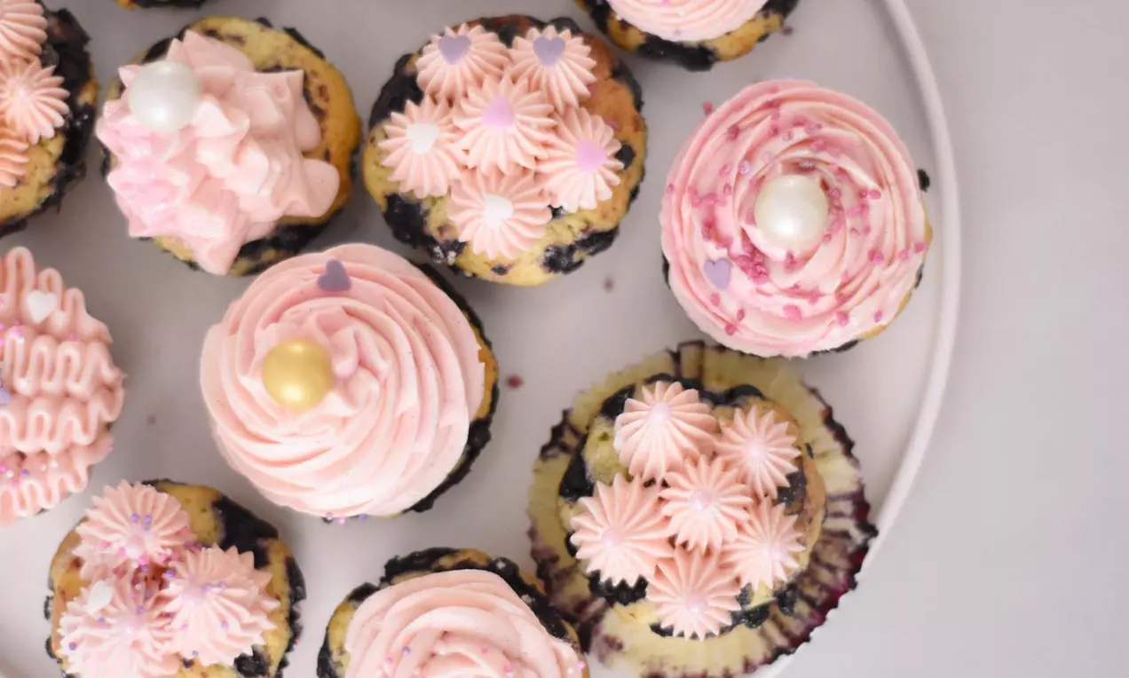 Muffins toppet med lyserød frosting i snoede mønstre, pyntet med perler og små blomster, anrettet på en hvid tallerken