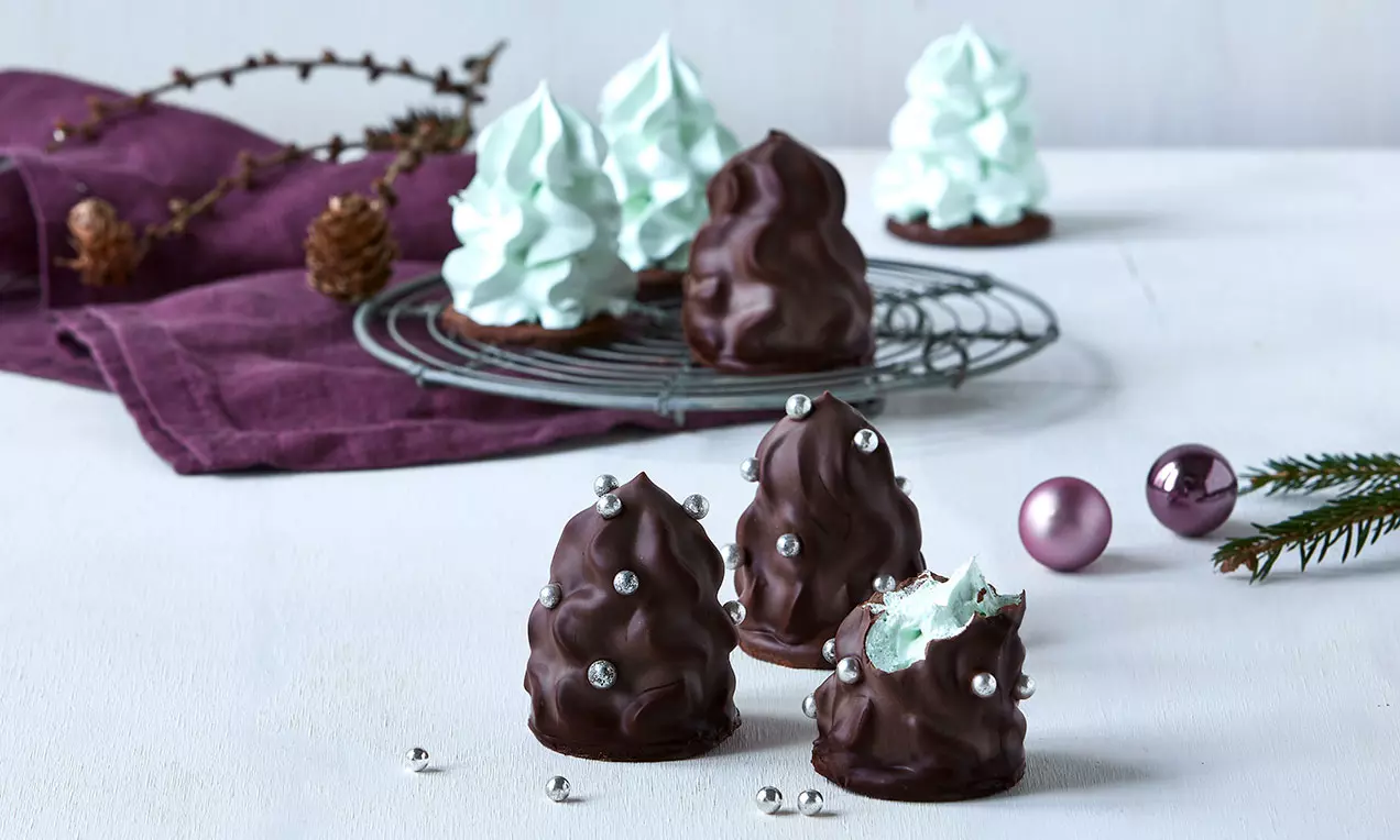 Chokoladeovertrukne flødeboller med lysegrønt skum toppet med sølvkugler på hvidt bord med lilla stof og pyntede grene
