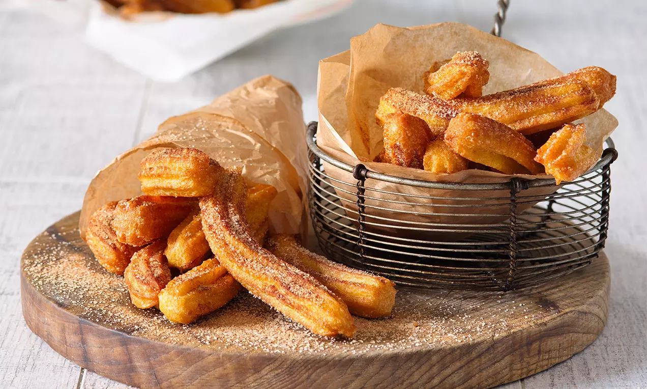 Gyldne churros med sprød overflade drysset med sukker, anrettet i brun papirpose og sort metalnetkurv på rustik træbræt