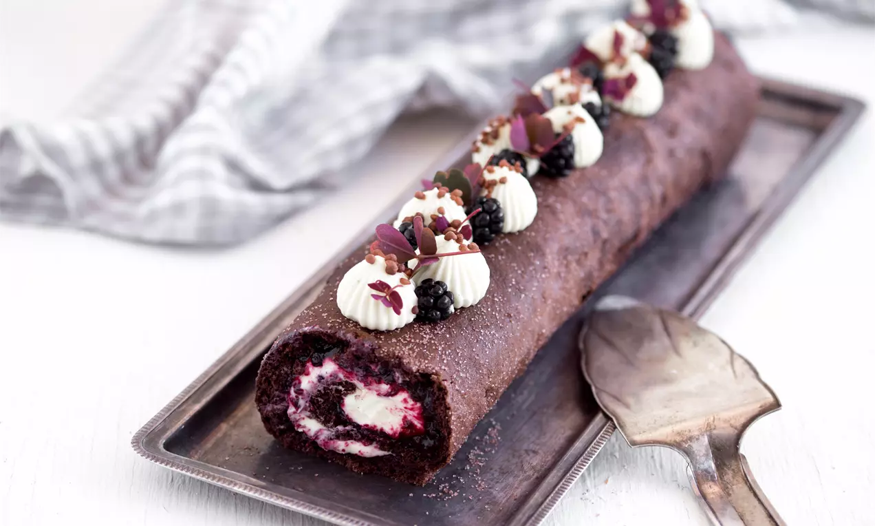 Aflang chokoladebrun roulade med hvid fyld og brombær, pyntet med fløde, blade og crumble på et sølvfad ved siden af en serverske