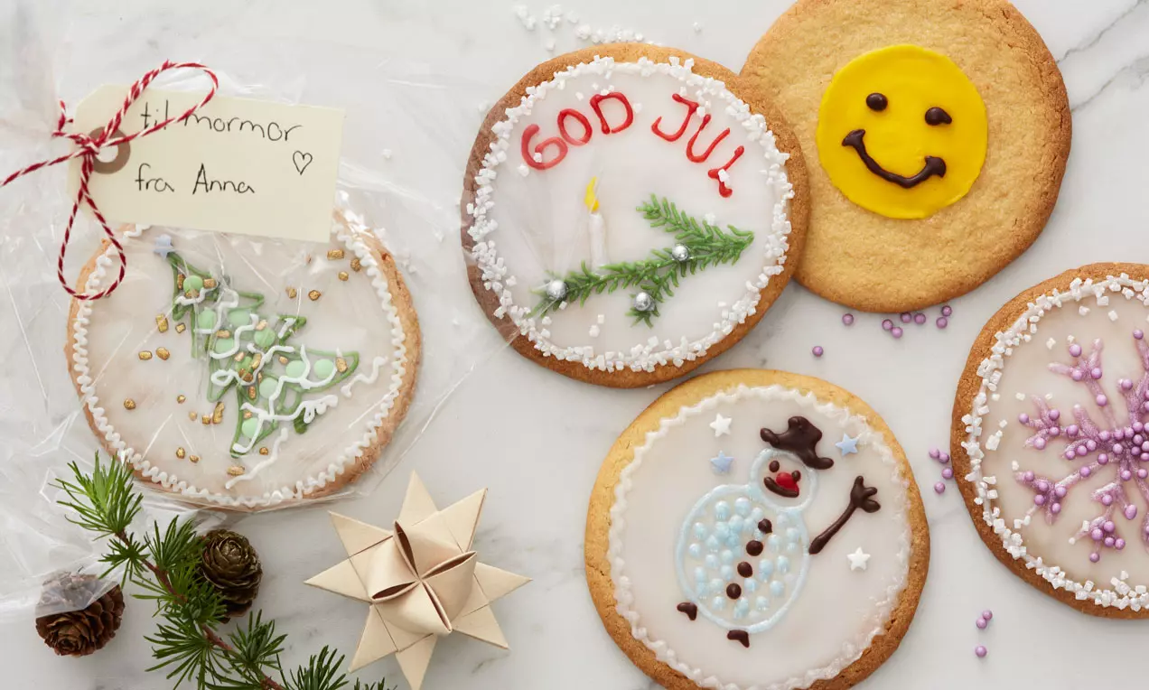 Store, flade småkager med glasurdekorationer som snefnug, juletræ og smil placeret på en hvid baggrund med grankogler og stjernedekoration