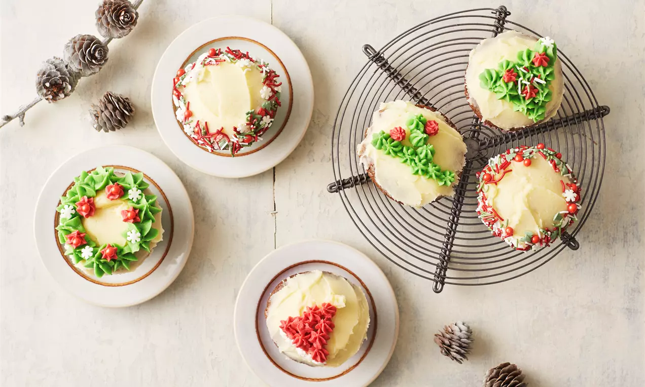Kryddercupcakes med lys glasur og røde, grønne og hvide juledekorationer anrettet på tallerkener og en sort bagerist med kogler som pynt