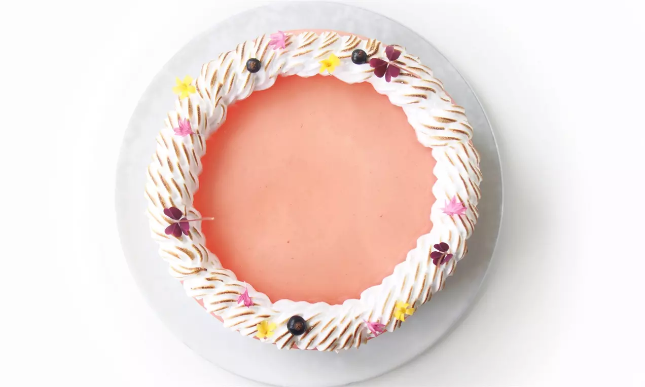 Vegetarisk cheesecake med blank karamel-overflade, hvid marengskrans med gyldne nuancer og små blomster på et hvidt kagefad