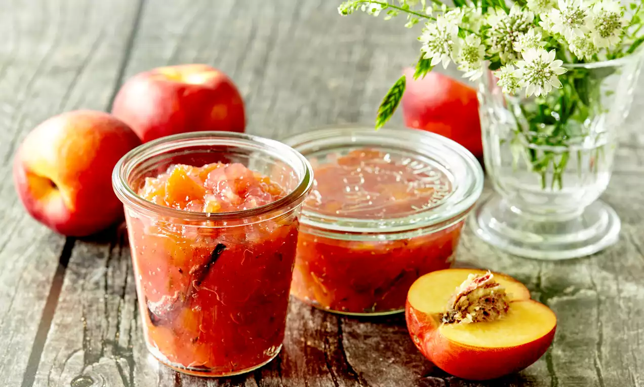 Gyldent orange ferskenmarmelade med synlige vaniljestænger i klare glas ved siden af halve ferskner og en vase med hvide blomster
