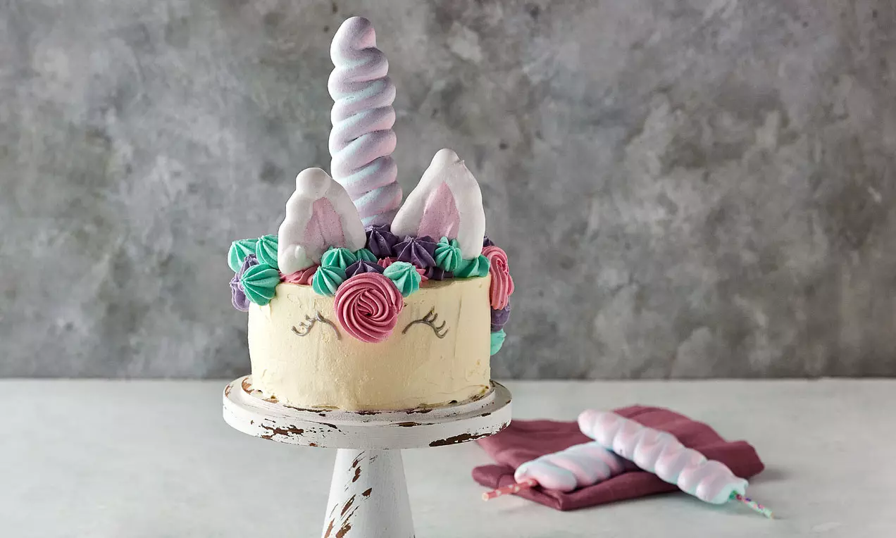 Lysegul kage med lilla, pink og blå marengsroser, toppet med snoet enhjørningehorn og marengsører på slidt hvidt kagefad