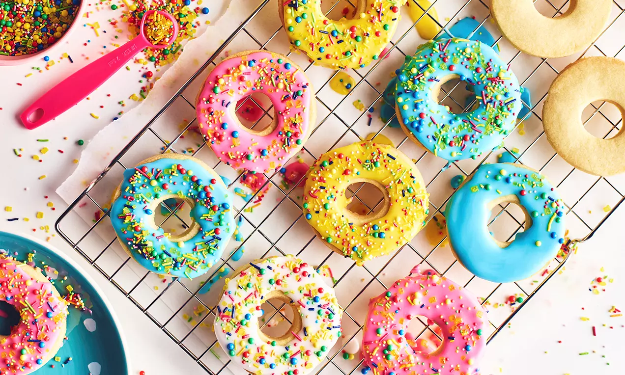 Småkager formet som doughnuts med farverig glasur og drysset med regnbue krymmel lagt på en metalrist rundt om spredte krymmel