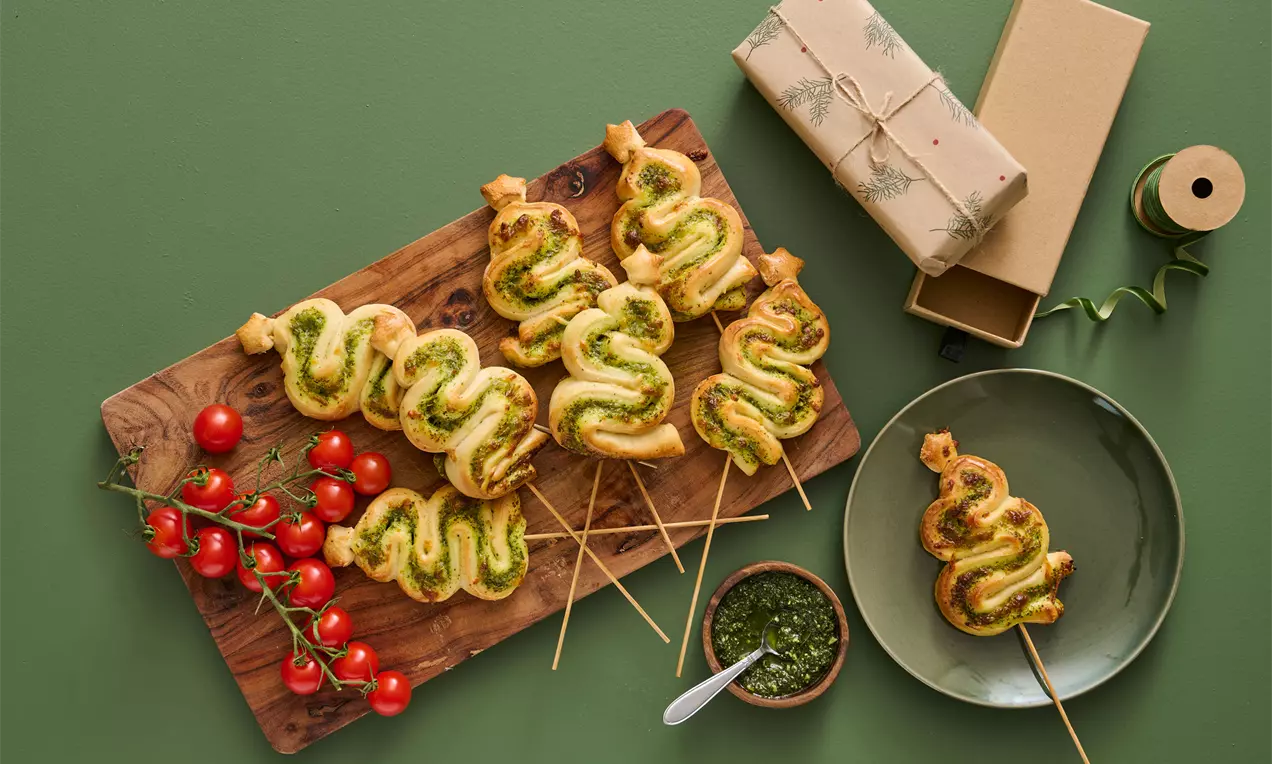 Gyldne juletræer af spiralformet pizzadej med grøn pesto på træspyd, anrettet på et skærebræt med røde tomater og skål med pesto
