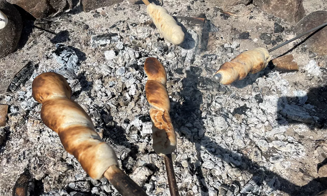 Snobrød med gyldne og lysebrune spiralformede skorper på pinde, ristet over glødende aske mellem spredte kul og sten
