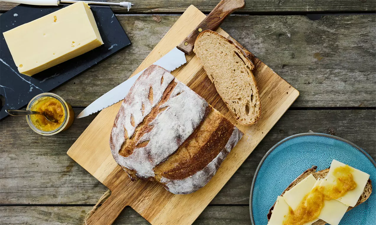 Surdejsbrød med gylden skorpe og melstrøg ligger på træbræt med brødkniv, skiver ved siden af og tallerken med brød, smør og marmelade