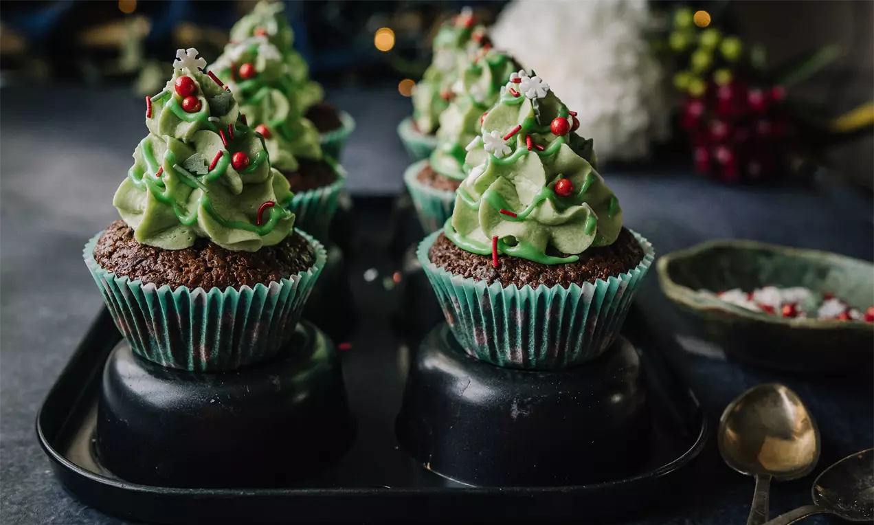 Cupcakes med mørkebrun bund i stribede blågrønne forme toppet med lysegrøn frosting, røde og hvide dekorationer på sort fad