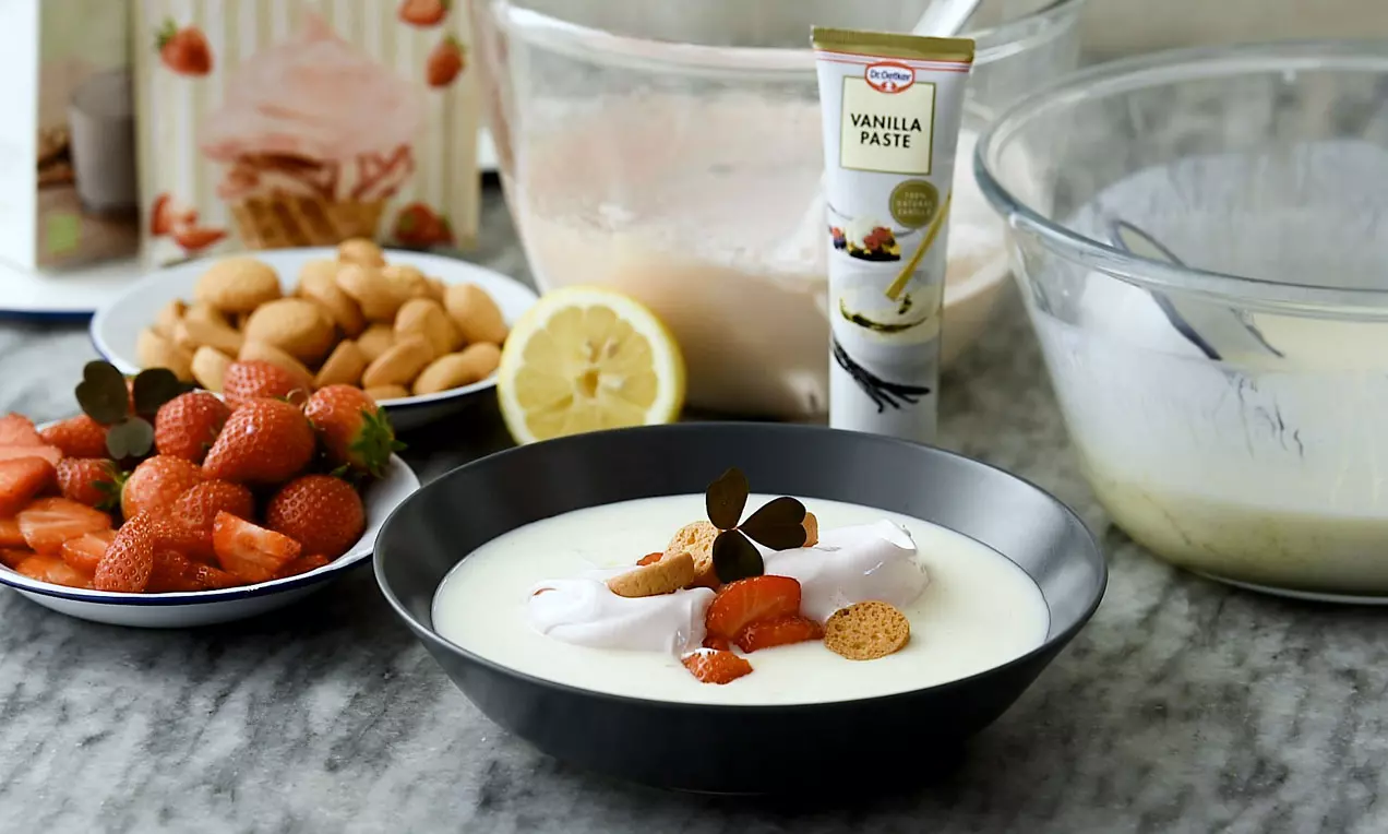 Hvid koldskål i sort skål med røde jordbær, små runde kammerjunkere og mørkegrønne urter på marmorbord med citron og vaniljecreme