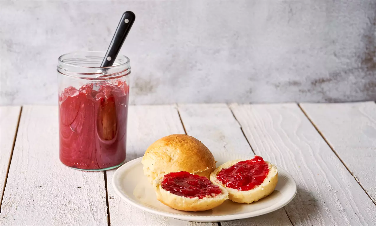 Rød skinnende marmelade med glat tekstur smurt på lyse boller, ved siden af et glas marmelade med metal-ske på træbord