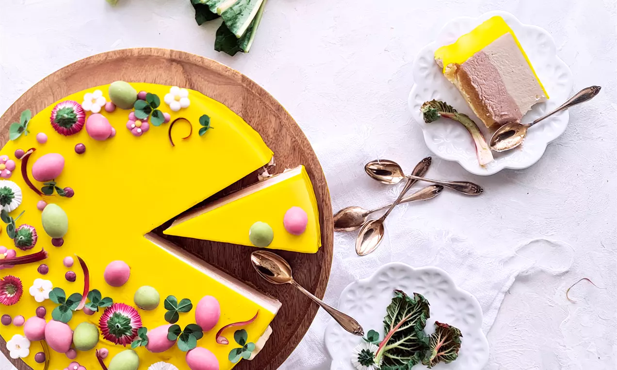 Rund påskekage med blank gul lemonglaze, pyntet med små farvede figurer og blomster på træfad, omgivet af hvide tallerkener og guldteskeer