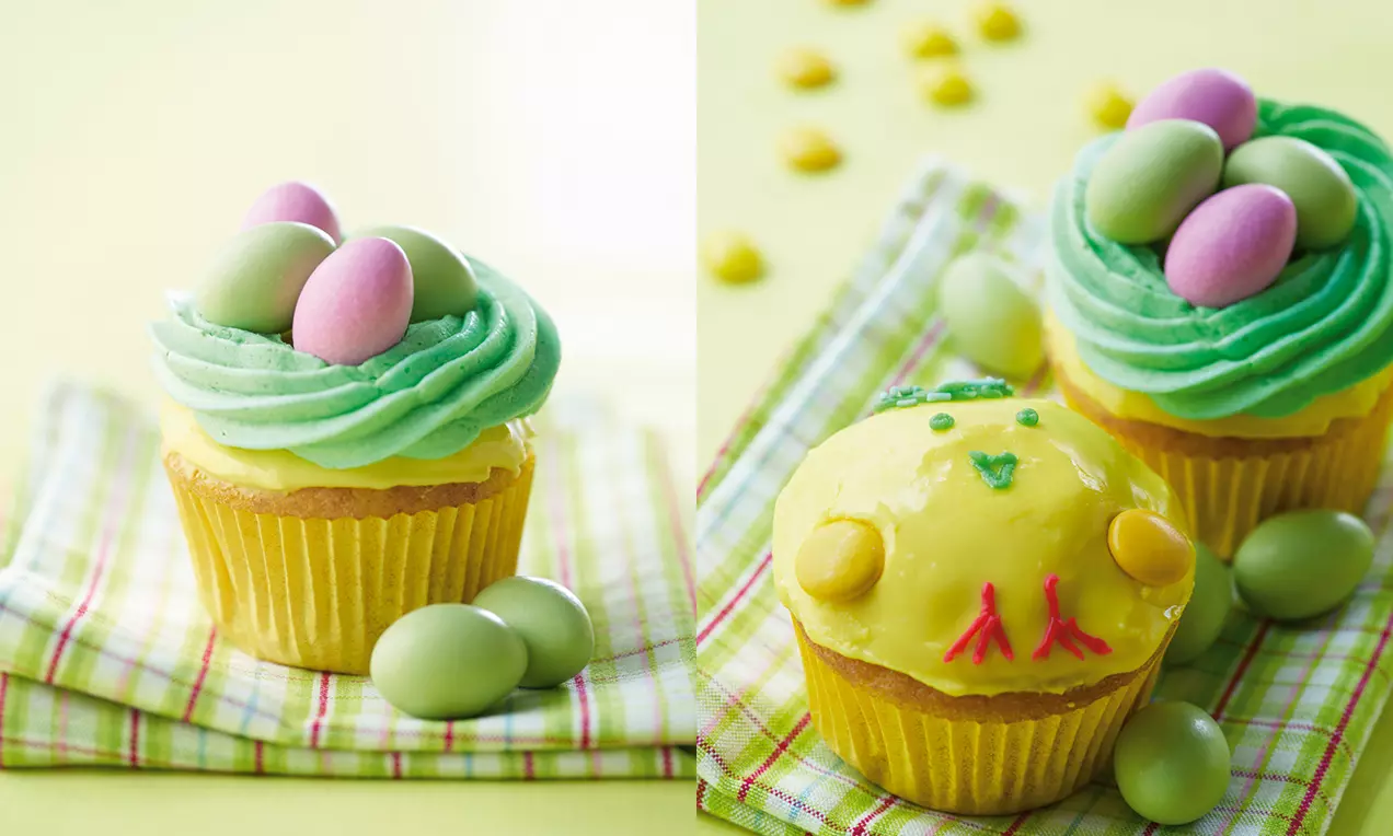 Påskecupcakes i gule forme med grøn frosting og farverige marcipanæg placeret på ternet stof mod en pastelgul baggrund