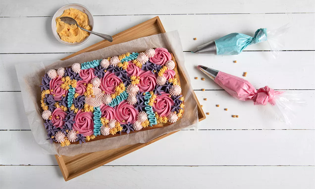 Rektangulær kage med farverige swirl-mønstre i lilla, pink, blå og gul frosting på bagepapir i en træbakke på hvidt bord