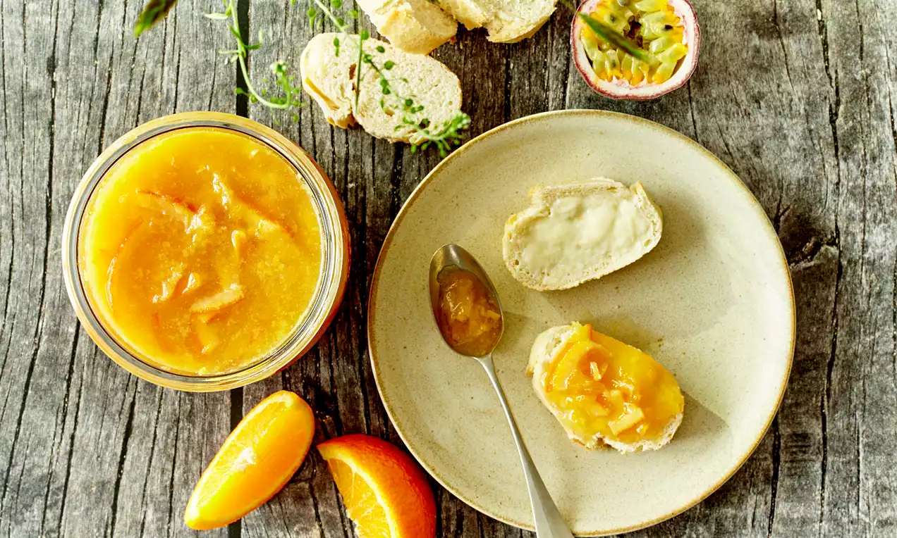 Appelsin-passionsfrugt marmelade med gyldentorange farve i et glas, smurt på lyse brødskiver ved siden af halve passionsfrugter