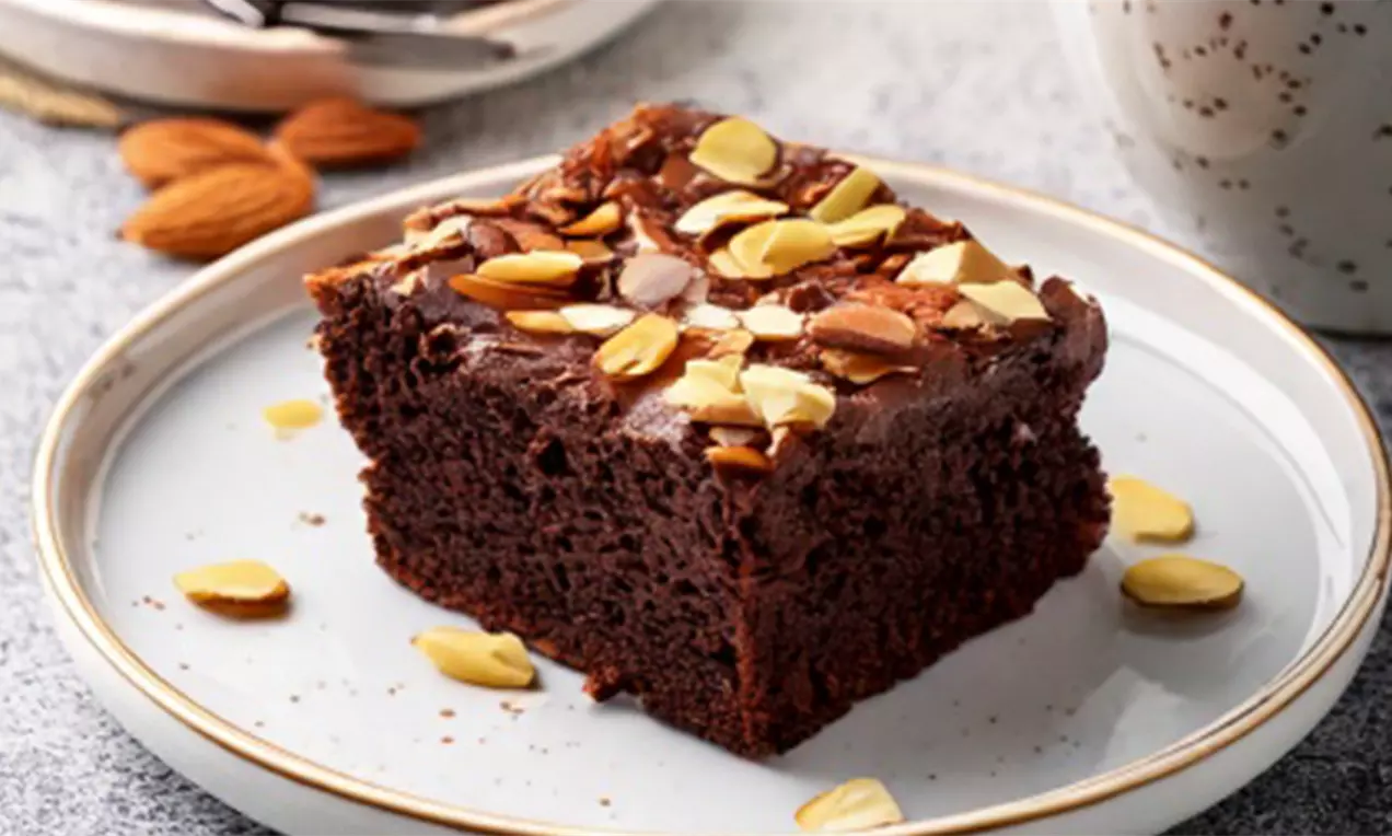 Firkantet, mørkebrun brownie med fugtig tekstur, drysset med lyse mandelflager på en lys tallerken med en kaffekop i baggrunden, AI-labeled image
