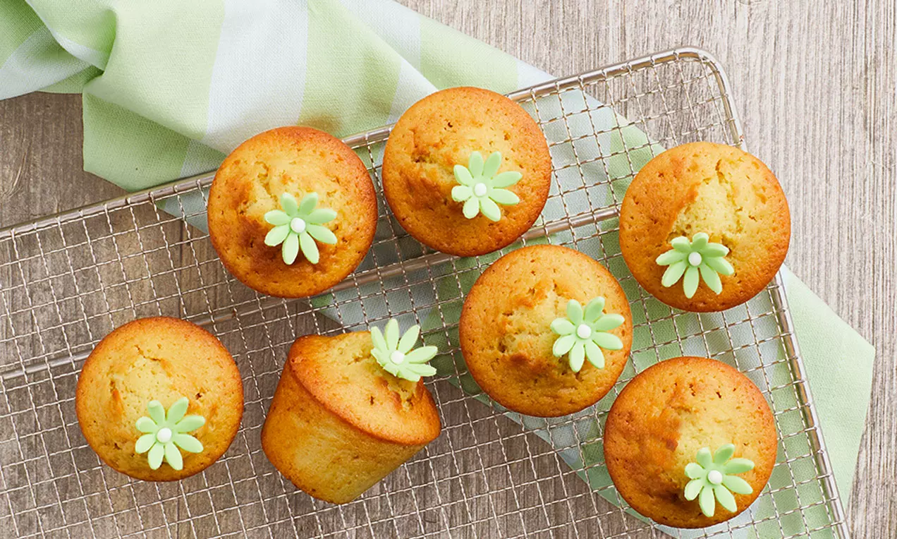 Gyldne muffins med let rustik top, pyntet med små lysegrønne fondantblomster, placeret på en metalrist med lysegrøn stofdekoration