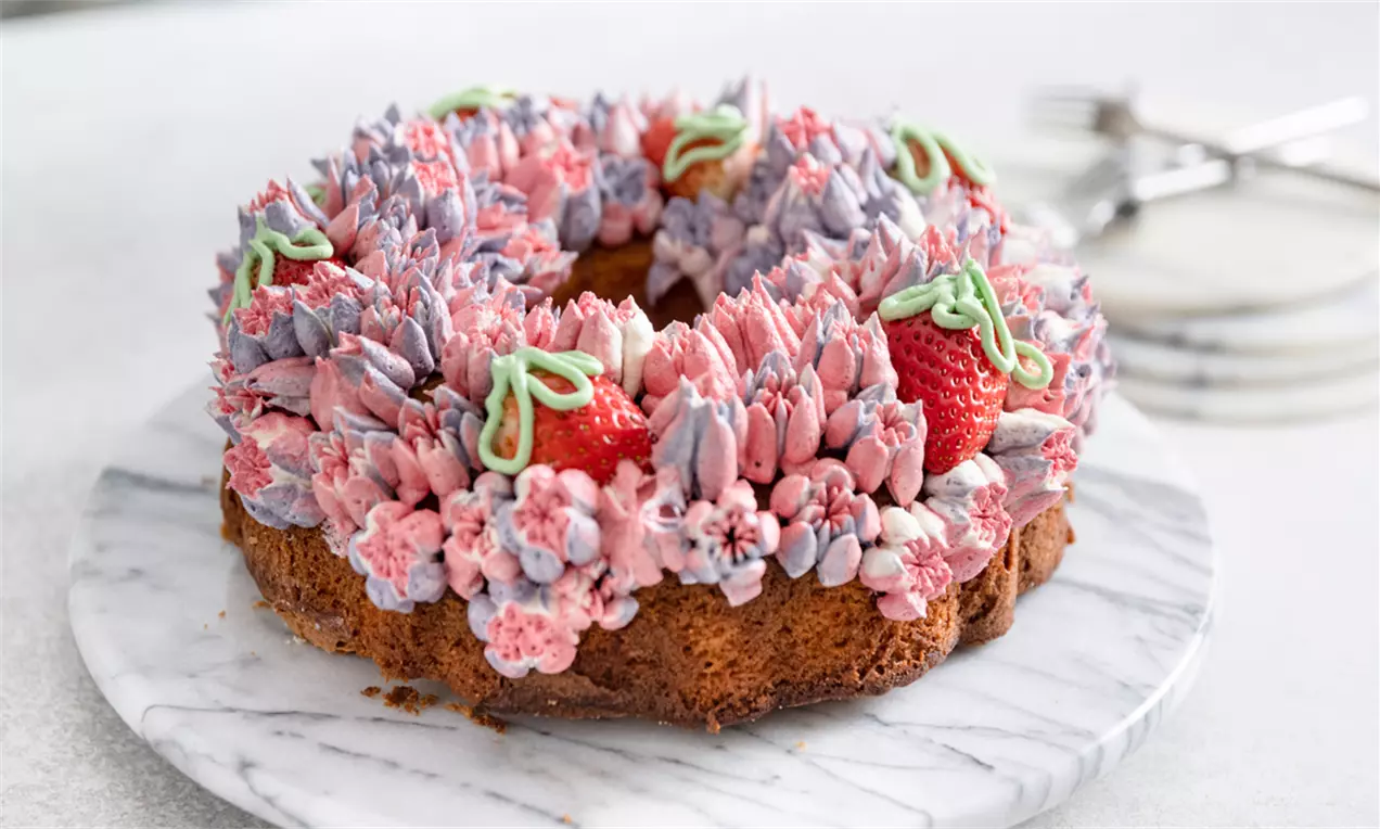 Rund lysebrun Bundtcake dækket med pink og lilla smørcremeblomster, friske røde jordbær og grønne blade på hvid marmorplade