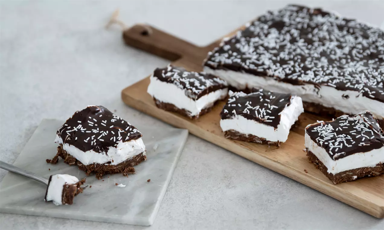 Firkantede stykker af flødebollekage med skummende hvid fyld og blank, mørk chokoladeoverflade drysset med kokos på træfad og marmorplade