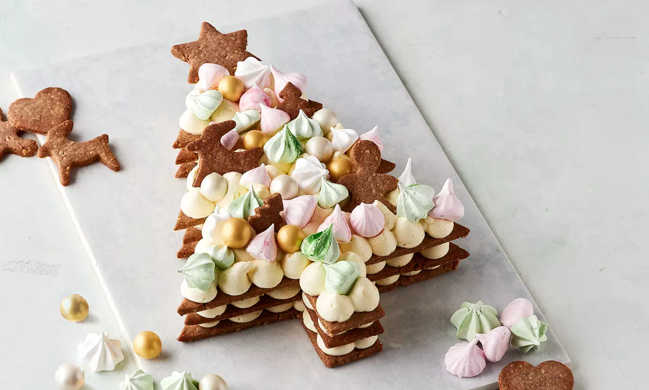 Juletræskage af brunlig sprød dej lagdelt med hvide creme-toppe og dekoreret med pastelfarvede marengs og gyldne kugler på hvidt fad